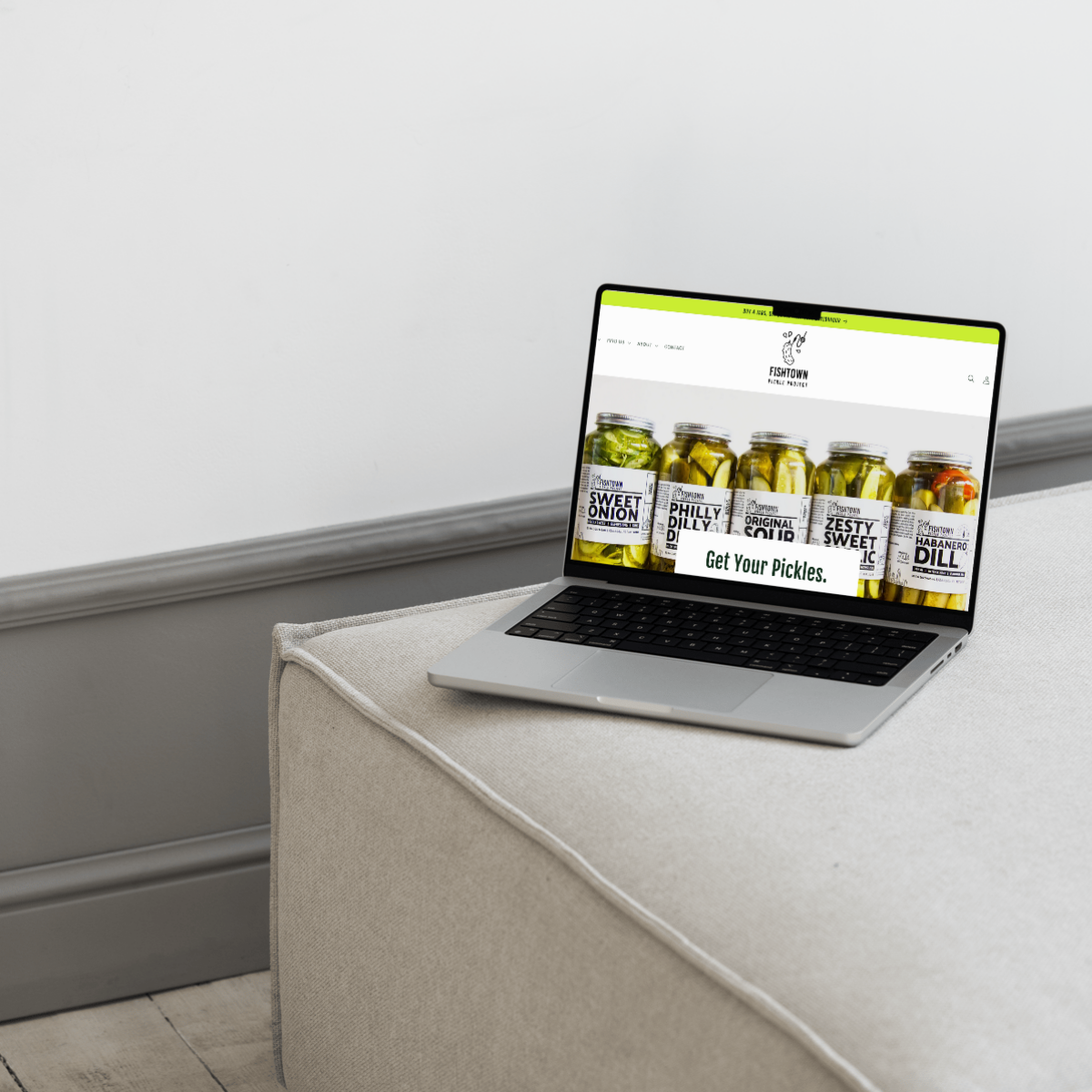  A laptop sitting on a neutral-colored ottoman displays the Fishtown Pickle Project e-commerce website. The homepage features a high-quality image of various pickled products with bold branding and a call-to-action button. This setup showcases a well-designed digital storefront for a specialty food brand.