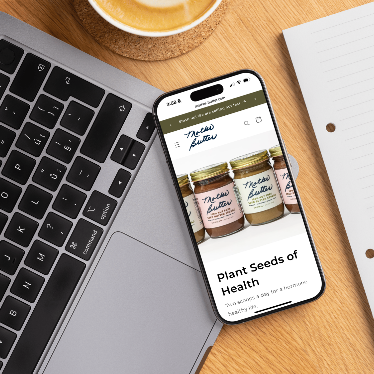 Smartphone displaying the Mother Butter ecommerce website, featuring plant-based nut butters with a health-conscious focus. The phone rests on a laptop near a cup of coffee, illustrating seamless online shopping for wellness brands. Ideal mobile website design for food startups and DTC brands.