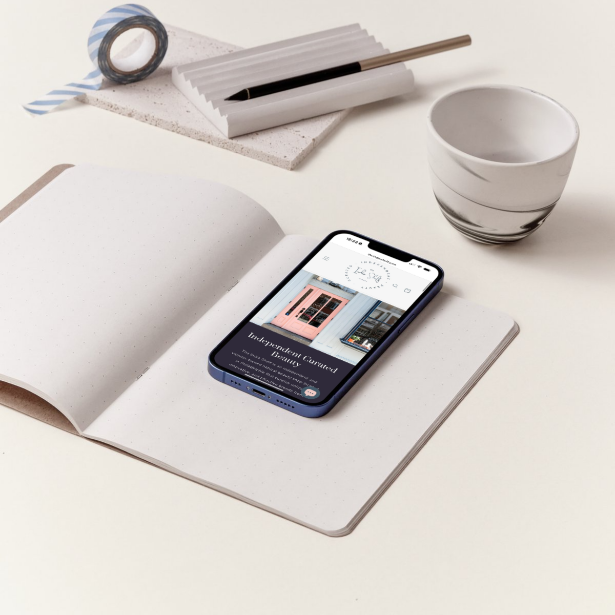 Smartphone showcasing The Indie Shelf’s ecommerce beauty store website, an independent Philadelphia-based shop. The phone is on an open notebook with a pen and coffee cup nearby, representing professional website design and email marketing for small businesses.